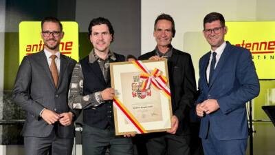 Vier Männer in Anzügen präsentieren eine gerahmte Urkunde mit Wappen und rot-weiß-rotem Band.