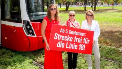 Drei Frauen halten ein rotes Plakat vor einer Straßenbahn der Linie 18.
