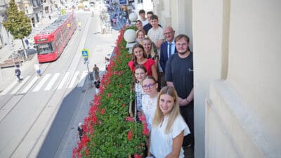 Gruppe von Lehrlingen und Begleitern auf Balkon mit roten Geranien und Blick auf Straße mit Straßenbahn