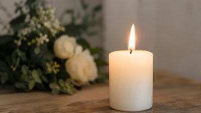 Eine brennende weiße Kerze auf einem Holztisch mit weißen Rosen und grünem Laub im Hintergrund.