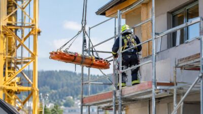 Feuerwehrmann in Schutzausrüstung auf einem Baugerüst neben einer hängenden orangefarbenen Rettungstrage