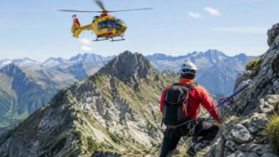 Bergsteiger in roter Jacke und Helm auf einem Felsen mit Rettungshubschrauber über den Bergen