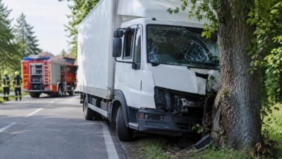 Weißer Klein-Lkw mit zerstörter Front und zersplitterter Windschutzscheibe lehnt nach Unfall an einem Baum neben der Straße