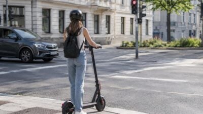 Eine Frau mit schwarzem Helm und Rucksack steht auf einem E-Scooter an einer roten Ampel.