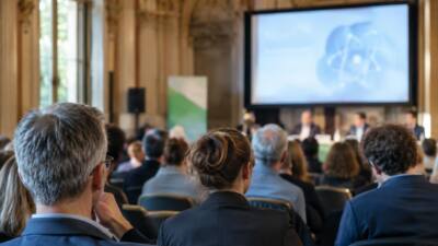 Publikum verfolgt eine Präsentation mit Atomsymbol in einem großen Veranstaltungssaal.