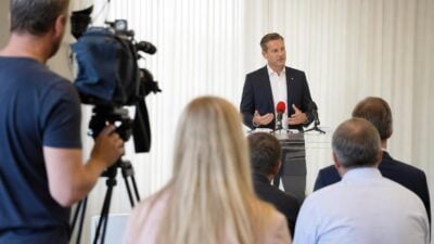 Ein Politiker in dunklem Anzug gestikuliert am Rednerpult mit Mikrofonen, im Vordergrund ein Kameramann und Zuschauer von hinten.
