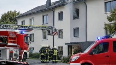 Feuerwehrleute in Schutzausrüstung stehen vor einem Gebäude während eine Drehleiter ausgefahren ist und Rauch aufsteigt