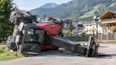 Ein auf die Seite gekippter roter Teleskoplader liegt auf einer asphaltierten Fläche vor Bergen und Häusern.
