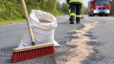 KI-Symbolbild mit Besen, Bindemittelsack und abgestreutem Straßenbelag; Feuerwehrkraft im Hintergrund.