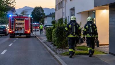 Zwei Feuerwehrleute in dunkler Schutzkleidung mit gelben Streifen gehen mit Geräten auf ein Haus zu.