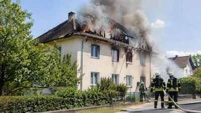 Feuerwehrleute in Schutzkleidung stehen vor einem brennenden Haus mit starker Rauchentwicklung am Dach.