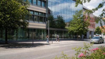 Modernes Bürogebäude mit Glasfassade, vor dem zwei Menschen auf dem Gehweg gehen.