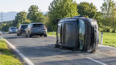 Ein schwarzer Pkw liegt nach einem Unfall auf der Seite am Straßenrand.