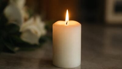 Eine weiße Kerze brennt mit einer Flamme auf einer grauen Tischplatte, unscharfe Blumen im Hintergrund.