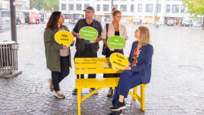 Vier Personen mit bunten Sprechblasen stehen und sitzen um eine gelbe Bank auf einem Platz.