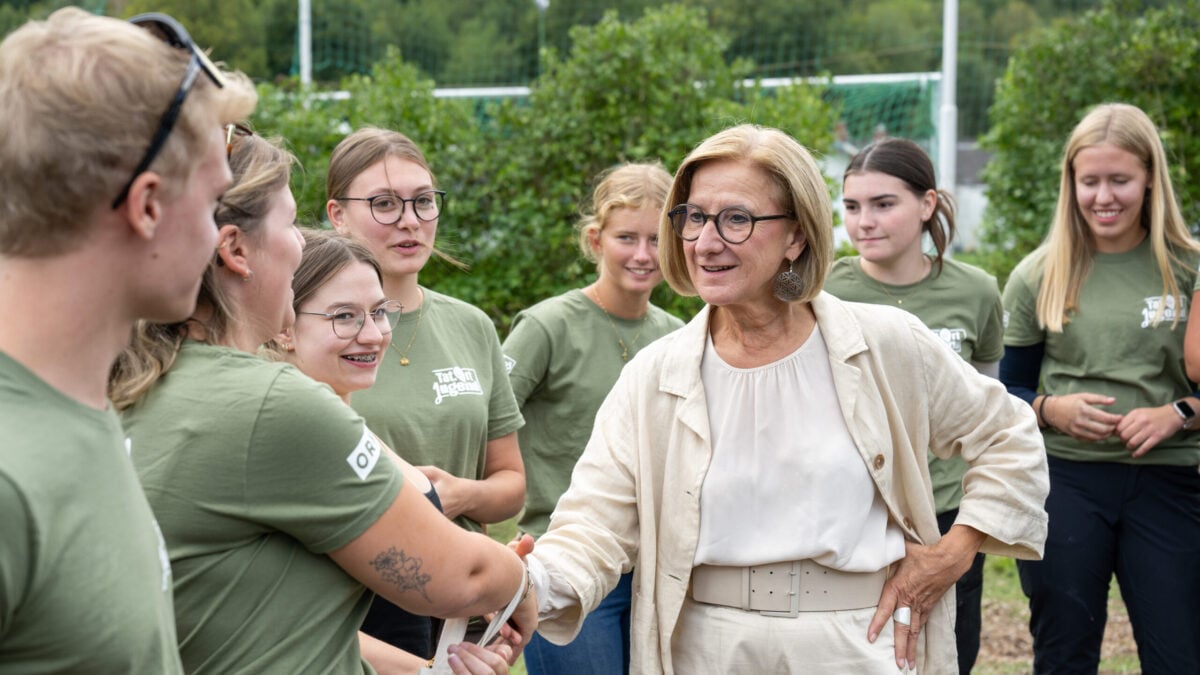 Ältere Frau mit Brille schüttelt einer jungen Frau in grünem T-Shirt die Hand vor einer Gruppe Jugendlicher.