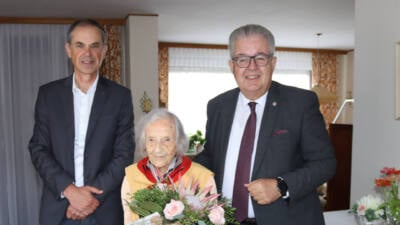 Eine ältere Frau mit Blumenstrauß steht zwischen zwei Männern in einem Wohnraum.