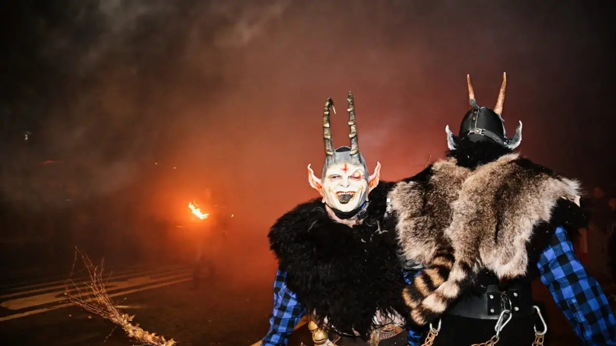 Zwei Krampusdarsteller in Hörnermasken und Fellkostümen vor rauchigem, orange beleuchtetem Hintergrund