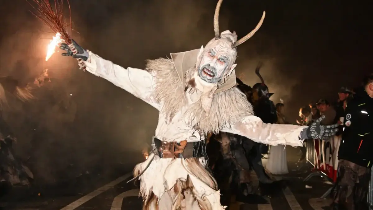 Eine gehörnte Krampusfigur in hellem Fellkostüm hält einen Funkenwedel in die Höhe.