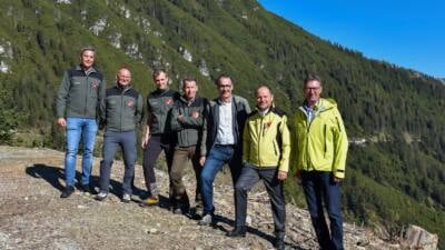 Menschen stehen auf einem kahlen Hang vor einem bewaldeten Tiroler Berghang.