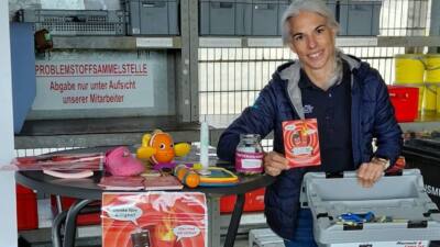 Eine Frau zeigt eine Akku-Infokarte neben Spielzeug und einer Sammelbox mit Batterien.