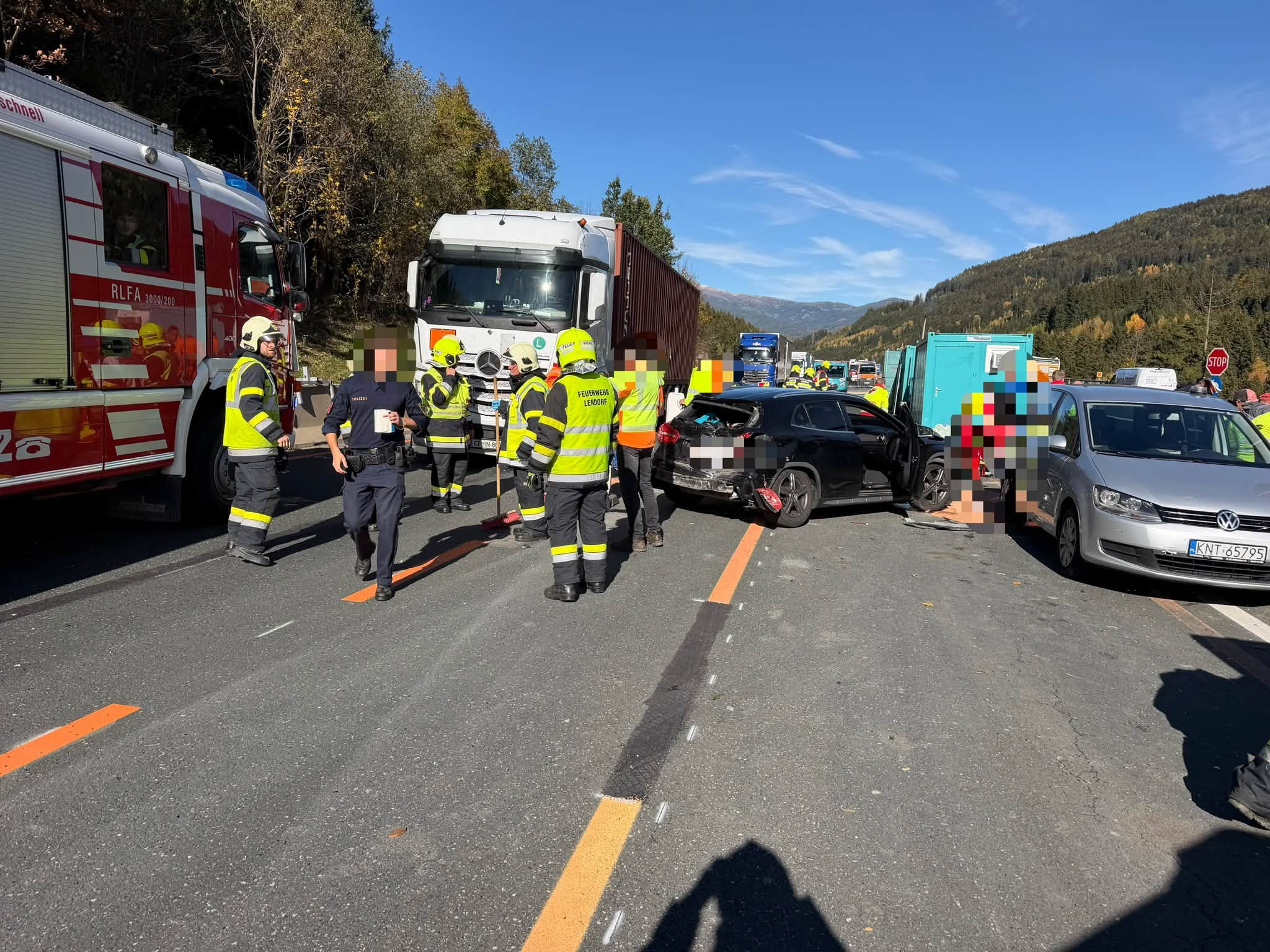 Feuerwehrleute und Einsatzkräfte stehen neben Lastwagen und beschädigten Autos auf einer Straße.