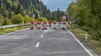 Rot-weiße Absperrgitter blockieren eine asphaltierte Bergstraße zwischen bewaldeten Hängen.