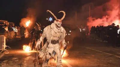 Eine Krampusfigur mit Hörnern und Fellkostüm steht nachts vor Zuschauern und Feuerschein.
