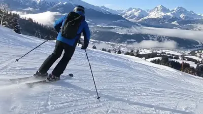 Eine Skifahrerin fährt auf einer verschneiten Piste vor einer alpinen Bergkulisse.