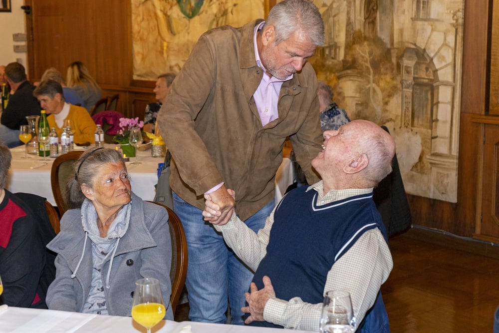 Ein Mann begrüßt einen sitzenden älteren Gast per Handschlag, daneben sitzt eine ältere Frau.