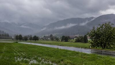KI-Symbolbild: Eine regennasse Straße vor grünen Wiesen und wolkenverhangenen Bergen.