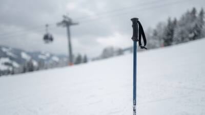 KI-generiertes Bild: Ein dunkler Skistock steckt im Schnee vor einer verschneiten Piste und einer unscharfen Seilbahn.