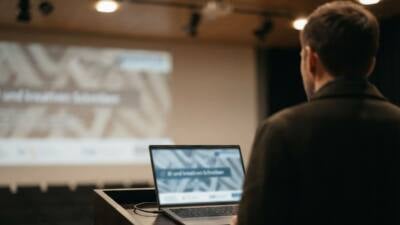 KI-generiertes Bild: Eine Person steht an einem Pult mit Laptop vor einer projizierten Präsentation.
