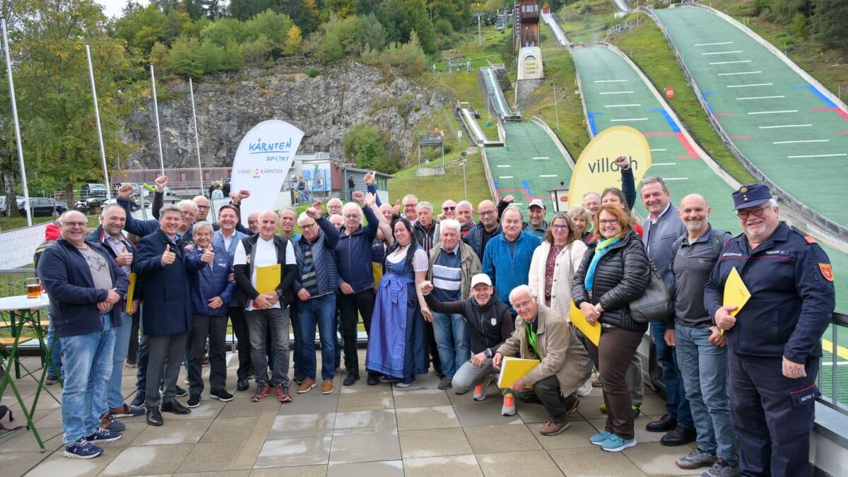 Gruppe von Erwachsenen vor Skisprungschanzen, einige halten gelbe Mappen.