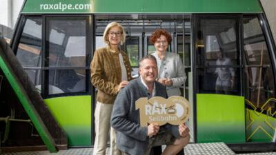 Vor einer grünen Seilbahnkabine präsentieren Personen ein Schild zum 100-jährigen Bestehen der Rax-Seilbahn.