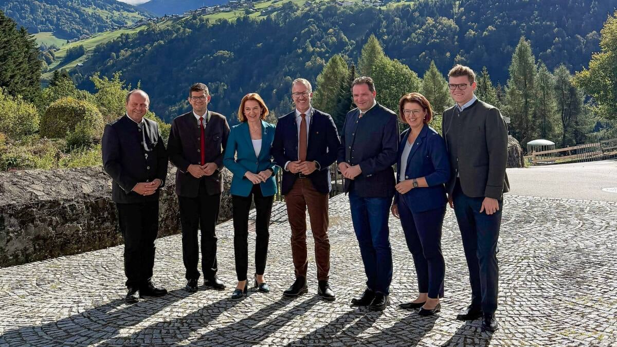 Eine Gruppe formell gekleideter Menschen steht vor einer bewaldeten Berglandschaft.