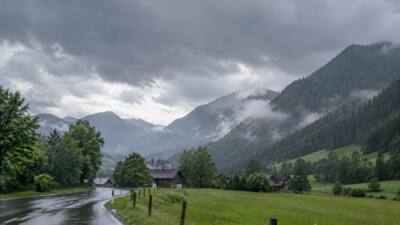 KI-Symbolbild: Eine nasse Straße führt durch ein grünes Alpental unter dunklen Wolken.