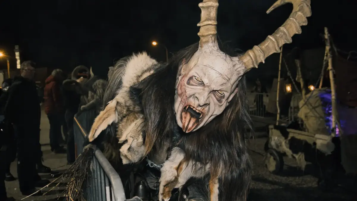 Ein Krampus in zotteligem Fell und großer Hörnermaske steht nachts an einer Absperrung.