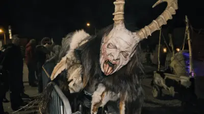 Ein Krampus in zotteligem Fell und großer Hörnermaske steht nachts an einer Absperrung.